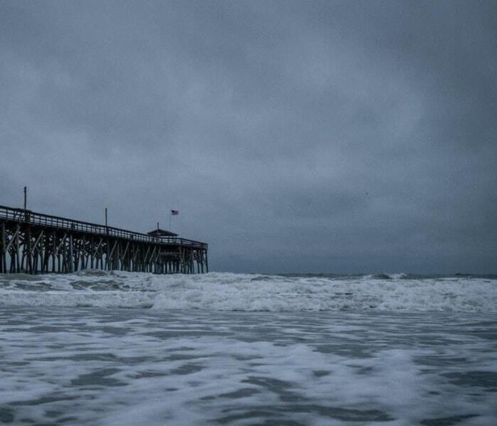 Ominous grey clouds hover over an old-looking oceanic pier. Dark, turbulent waves can be seen in the foreground.